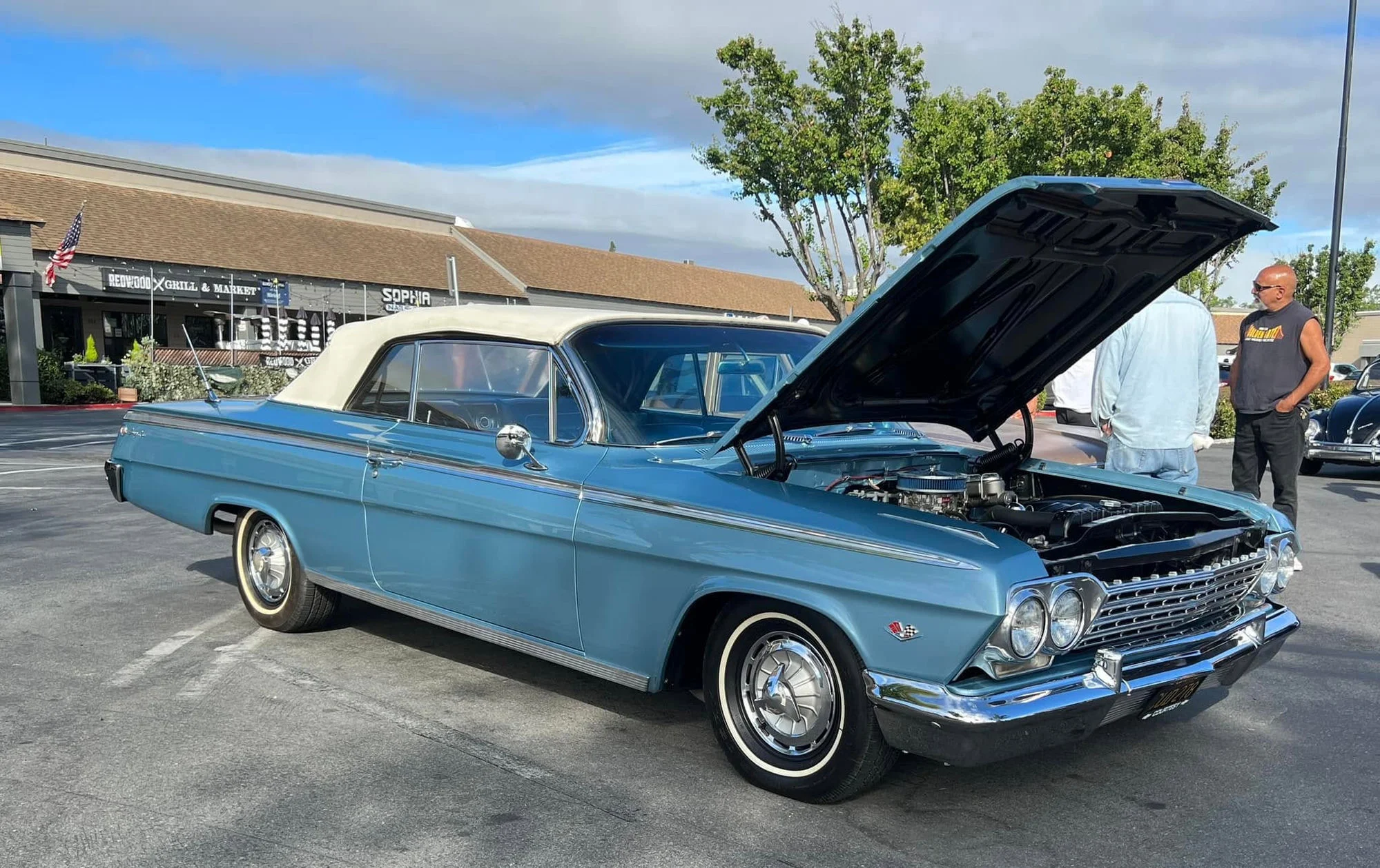 A restored teal 1962 Chevrolet Impala convertible with its hood open at Cars and Coffee Peninsula in the Woodside Plaza parking lot